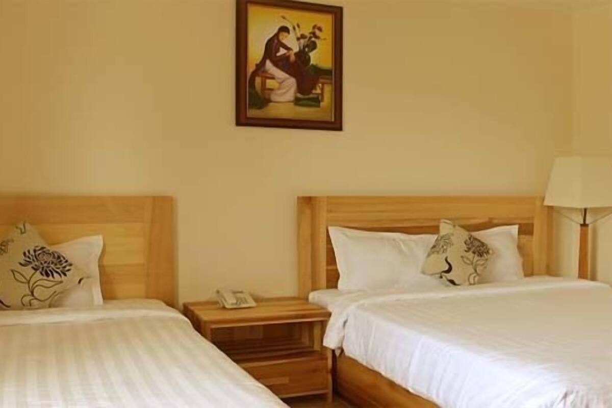 A hotel room with two single beds, wooden headboards, bedside tables, a lamp, and a painting on the wall.