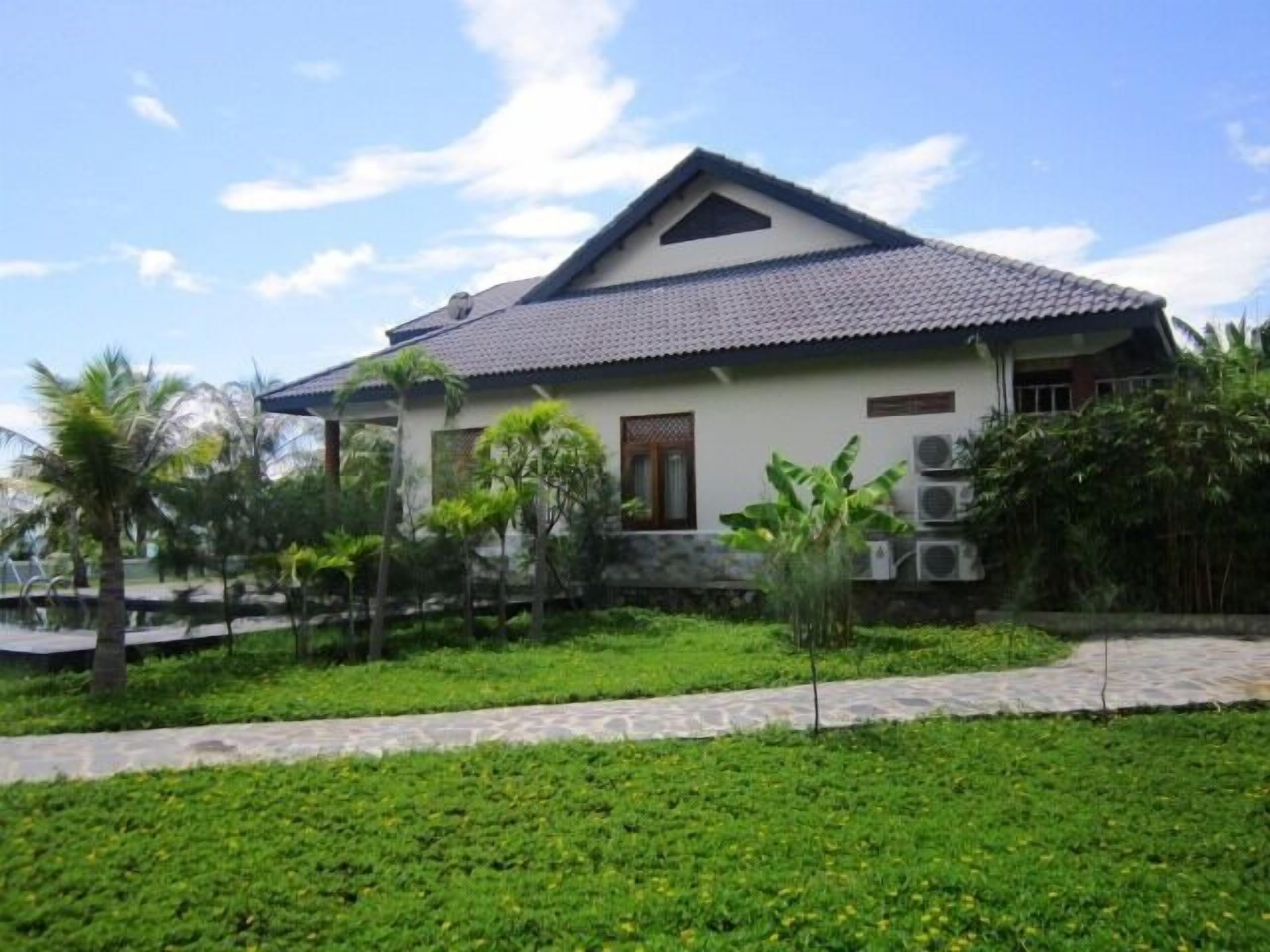 A single-story house with a tiled roof, surrounded by lush greenery and palm trees.