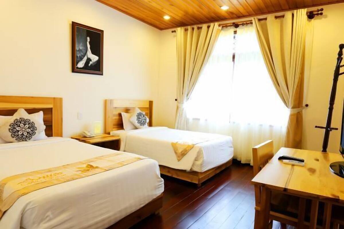 A hotel room with two beds, wooden flooring, a desk with a chair, and a window with curtains.