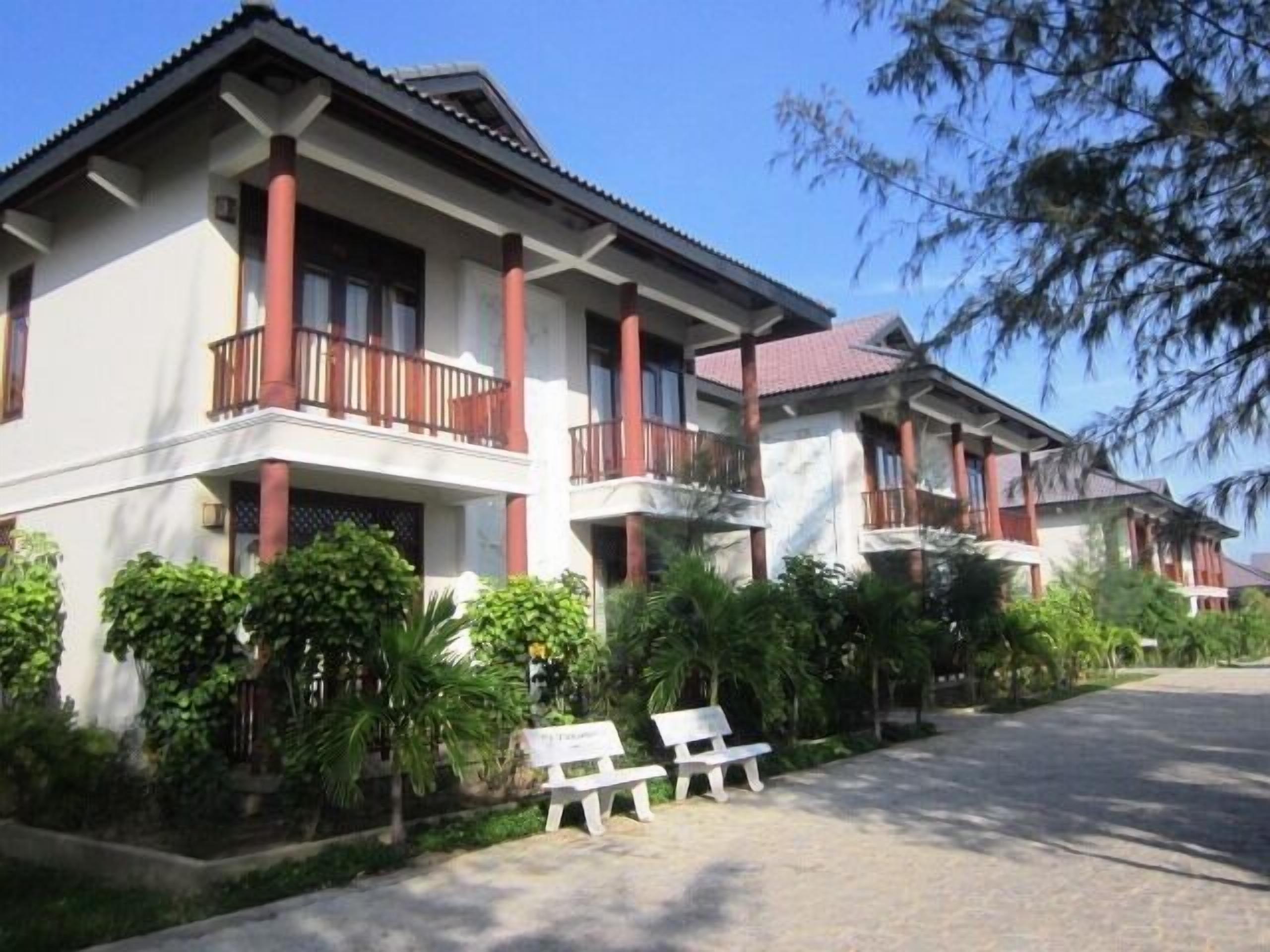 A row of two-story buildings with balconies, surrounded by greenery and a paved driveway.