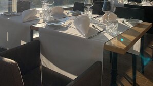 Un espace repas avec des tables préparées pour deux personnes, de grandes fenêtres offrant une vue sur les montagnes et une lumière vive qui filtre à travers.