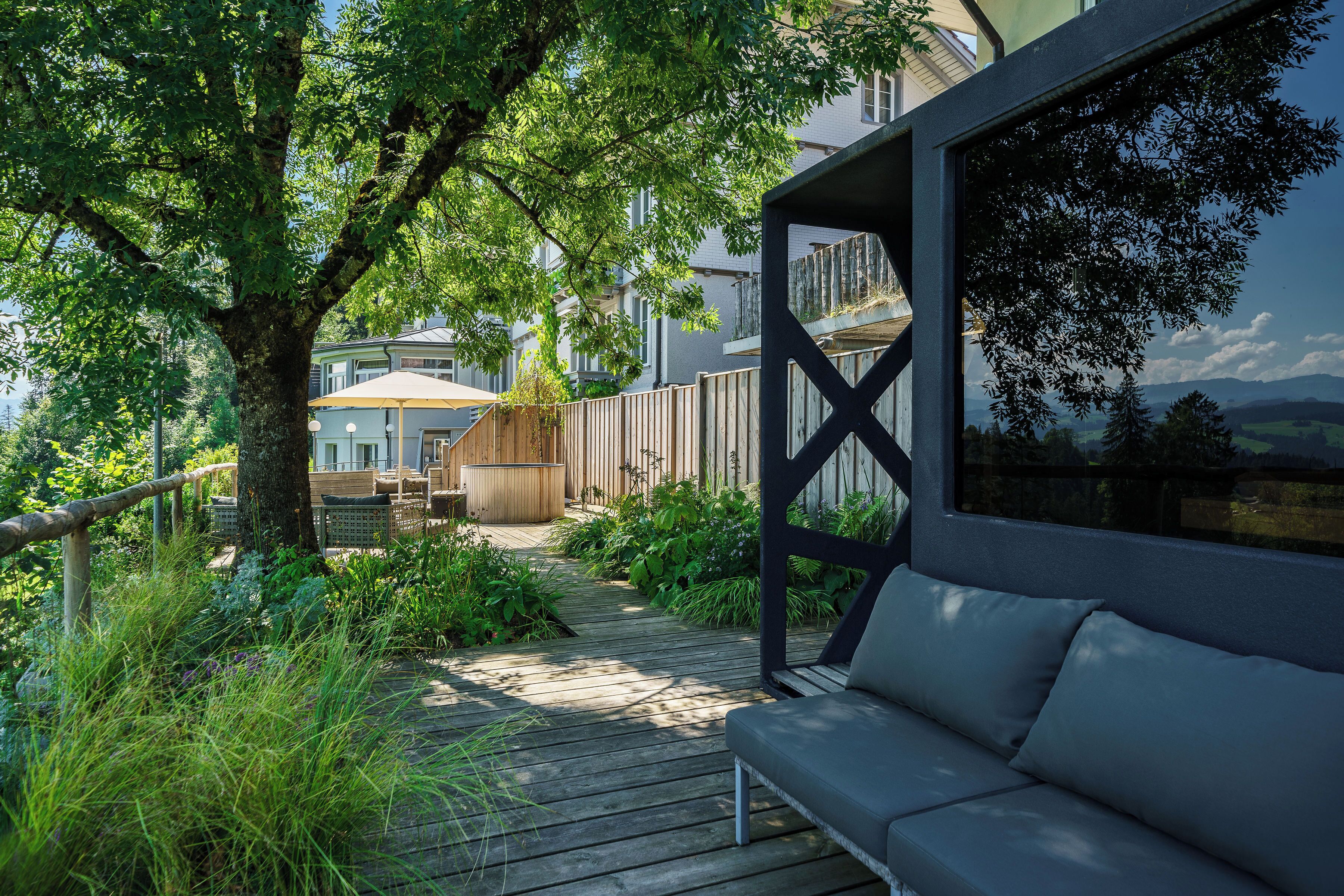 A wooden deck with a sofa, surrounded by lush greenery and a wooden fence.