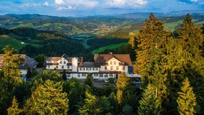 Un grand immeuble à étages, doté d’une façade symétrique, entouré d’une forêt dense et bordé de collines douces à l’arrière-plan.