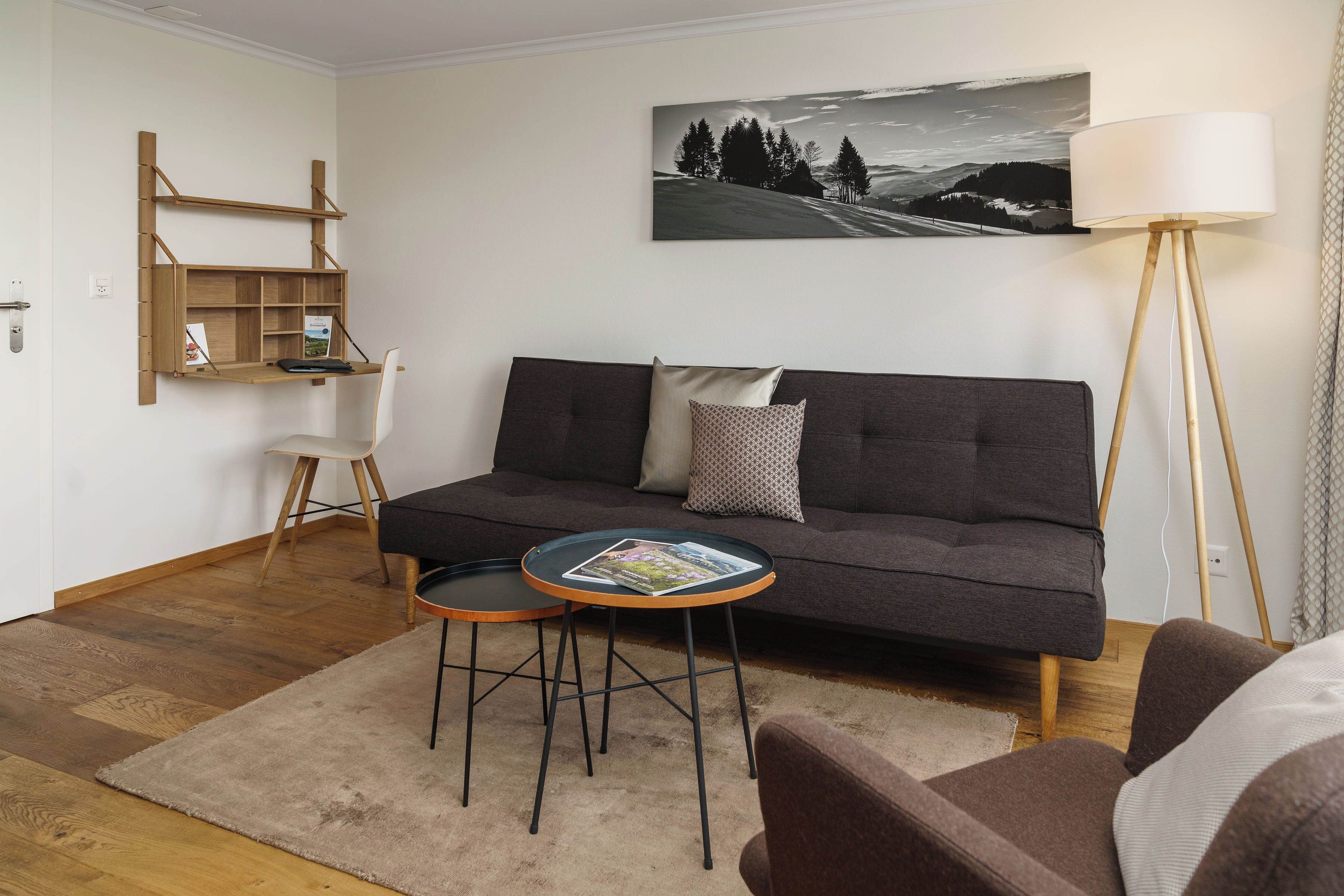 Un salon moderne avec un canapé gris foncé, une petite table ronde sur laquelle repose un magazine, une étagère en bois et un lampadaire.