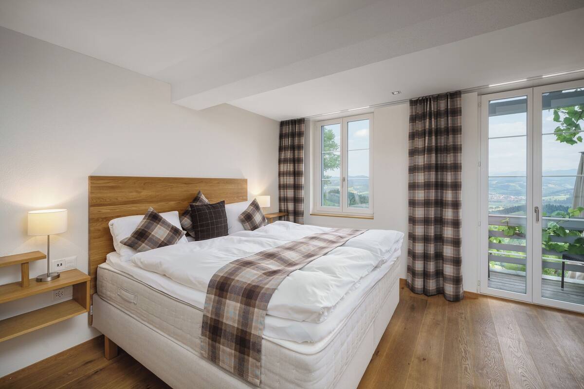 Un dormitorio con una cama grande, cabecera de madera, mesita de noche con lámpara y vista a través de puertas correderas de vidrio.