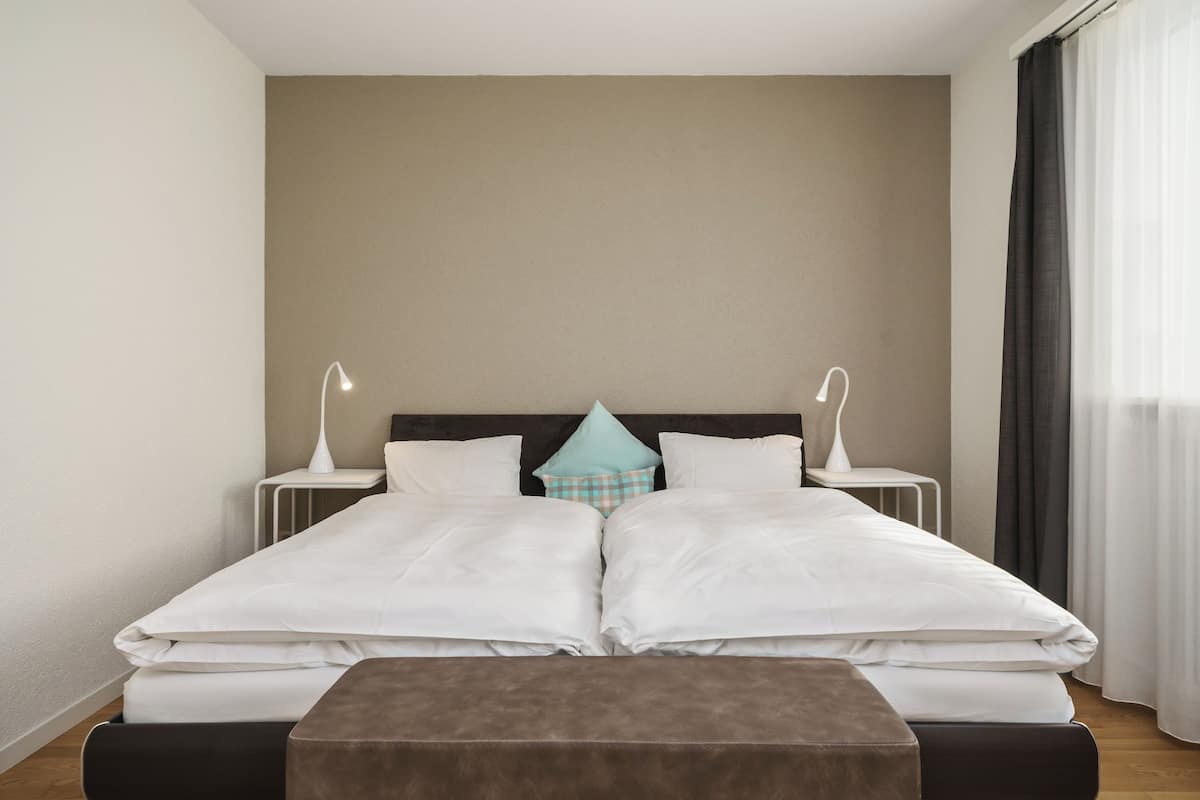 Cama doble con ropa de cama blanca, dos mesitas de noche con lámparas, un cojín de cuadros y un reposapiés marrón.