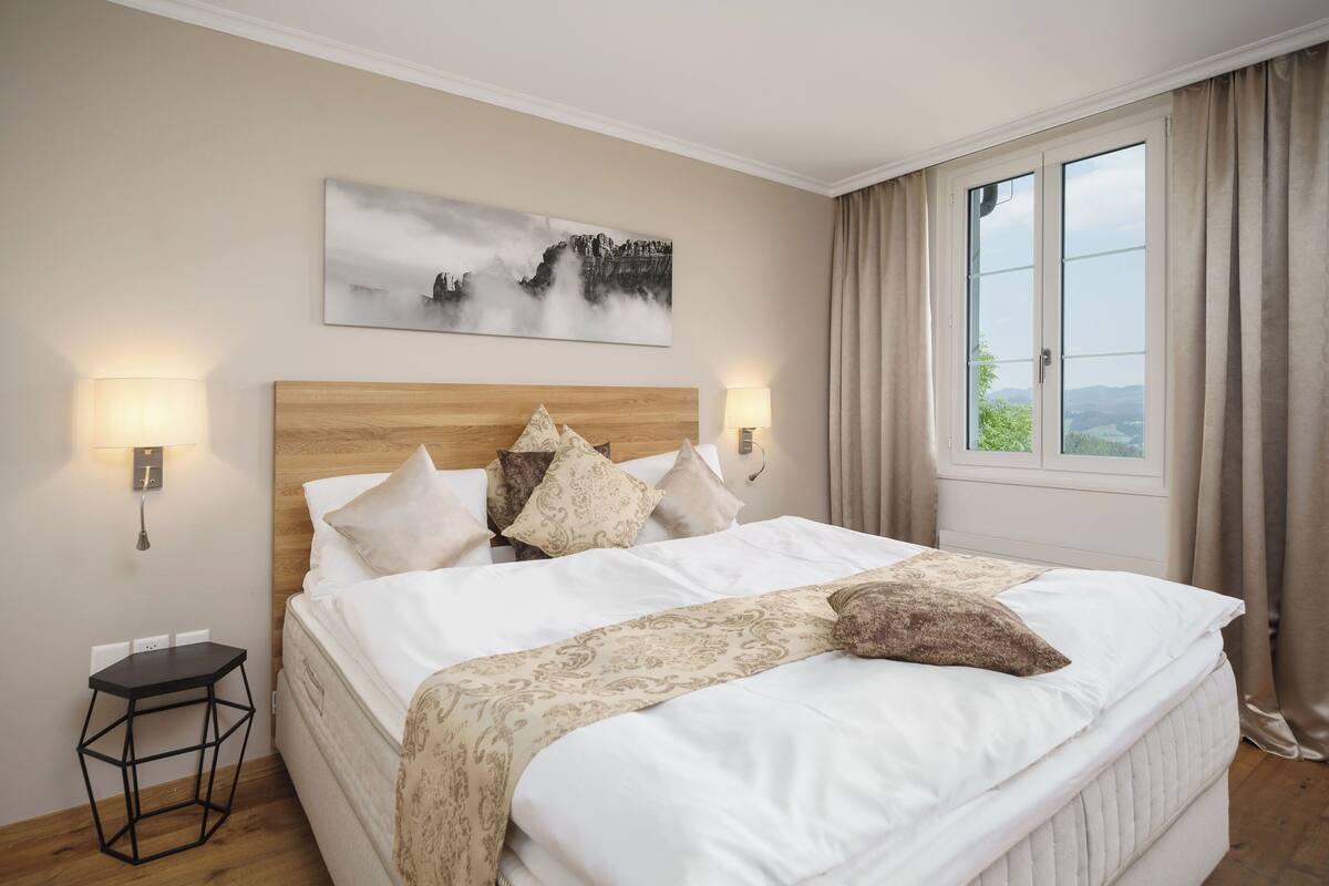 Una cama bien tendida con cabecera de madera, dos lámparas de noche, una mesita y un amplio ventanal con cortinas.