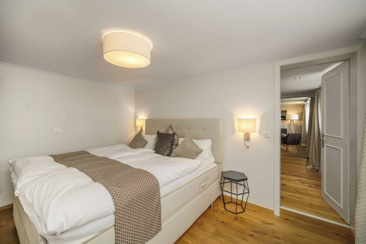 Un dormitorio con cama, lámparas de noche, una mesita de noche y una puerta que da a otra habitación.