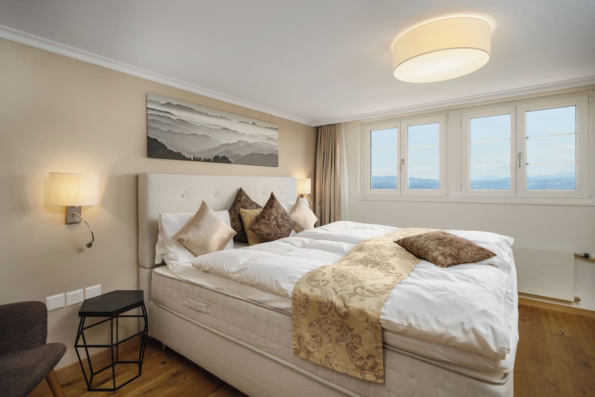 Un dormitorio con una cama grande, cabecera, mesita de noche, lámpara, un cuadro y una ventana con cortinas.