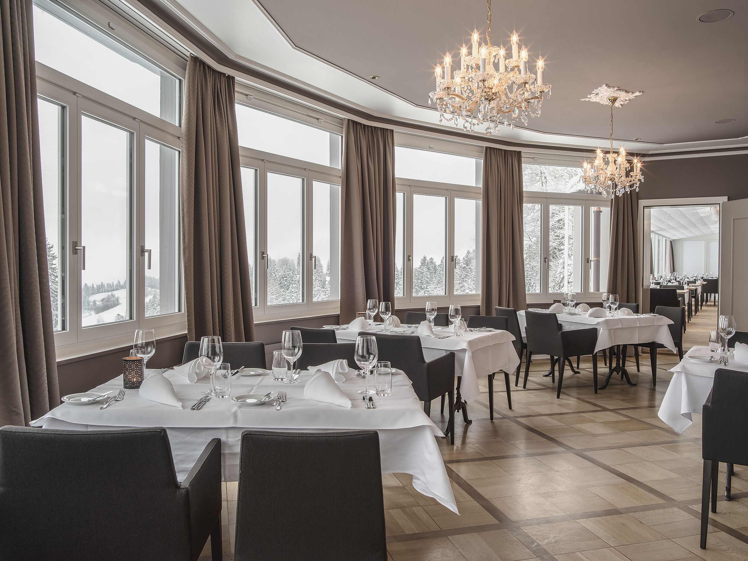 Un espace repas avec des tables dressées, de grandes fenêtres ornées de rideaux et un lustre.