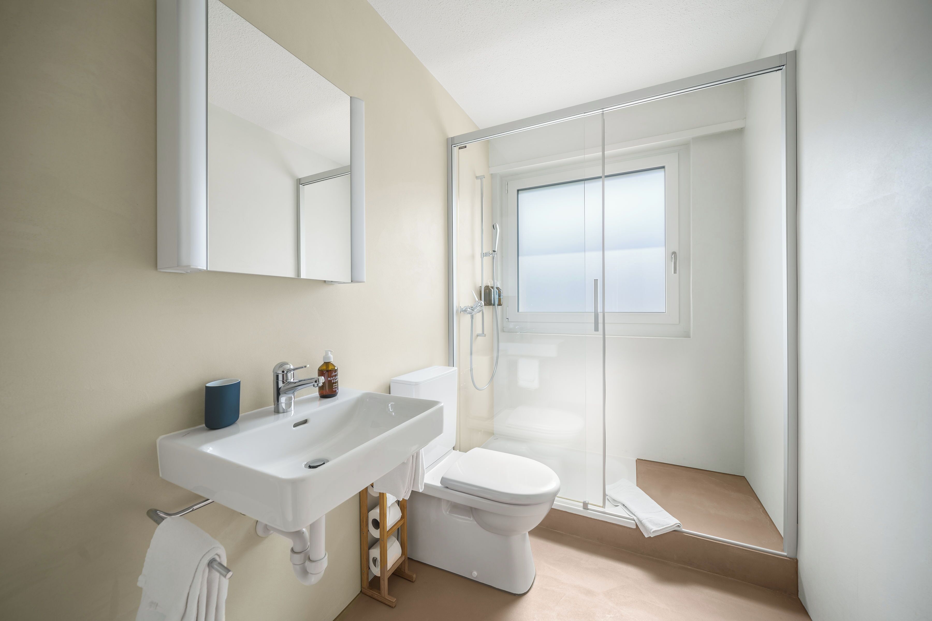 Un baño moderno con lavamanos blanco, espejo y retrete, además de cabina de ducha con mampara de vidrio.