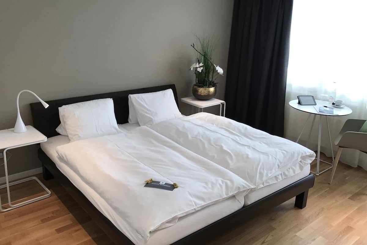Una cama bien hecha con sábanas blancas, una mesita de noche con lámpara, una pequeña mesa con una planta y una silla cerca de una ventana con cortinas.