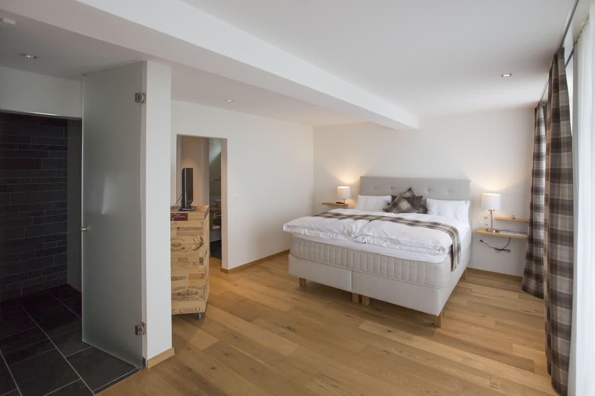 Un dormitorio con una cama grande, suelo de madera, una puerta de vidrio que da al baño y una pared de ladrillo.