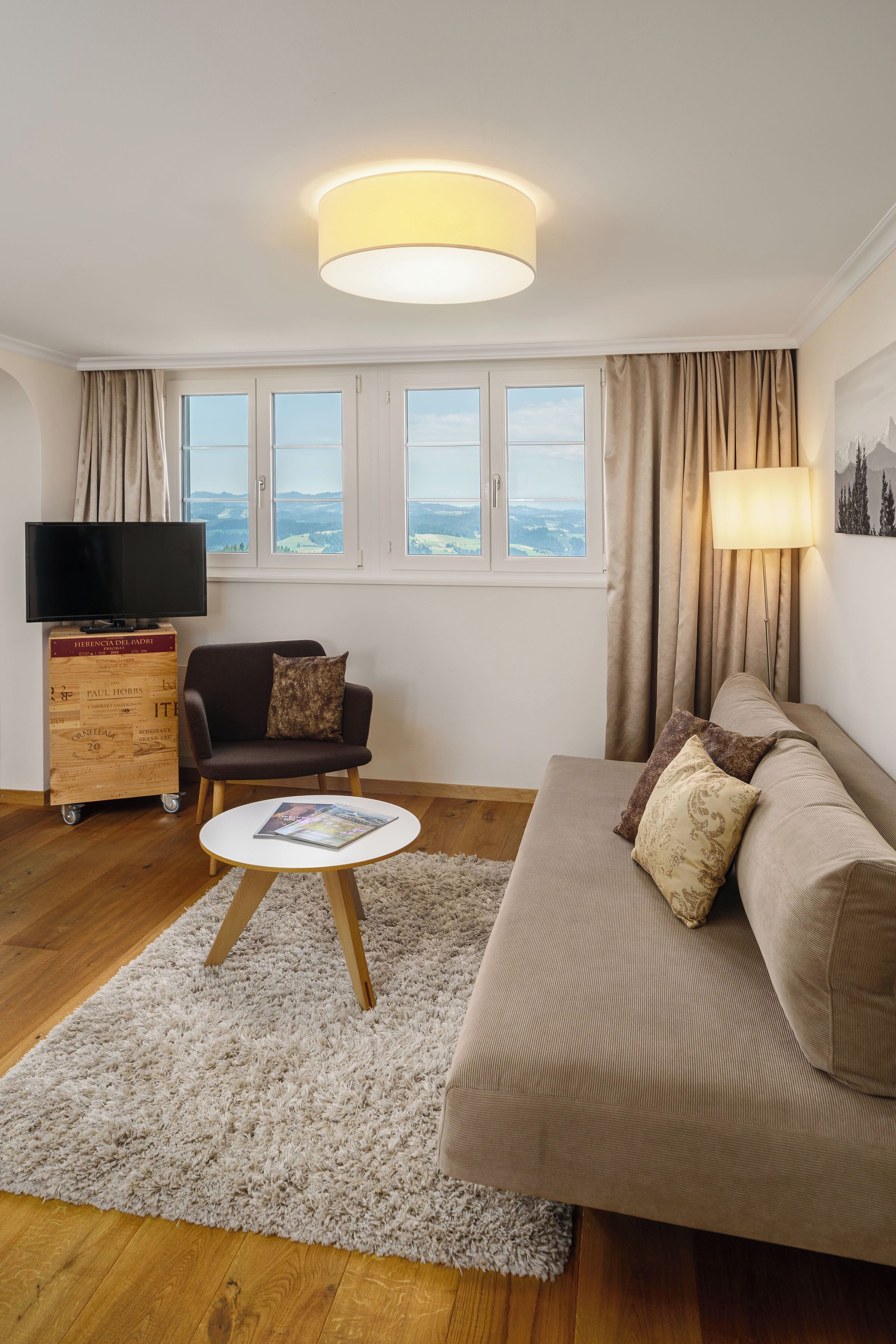 Uma sala de estar moderna com sofá, poltrona e uma pequena mesa de centro. Uma televisão está sobre um móvel de madeira, e há janelas com cortinas.