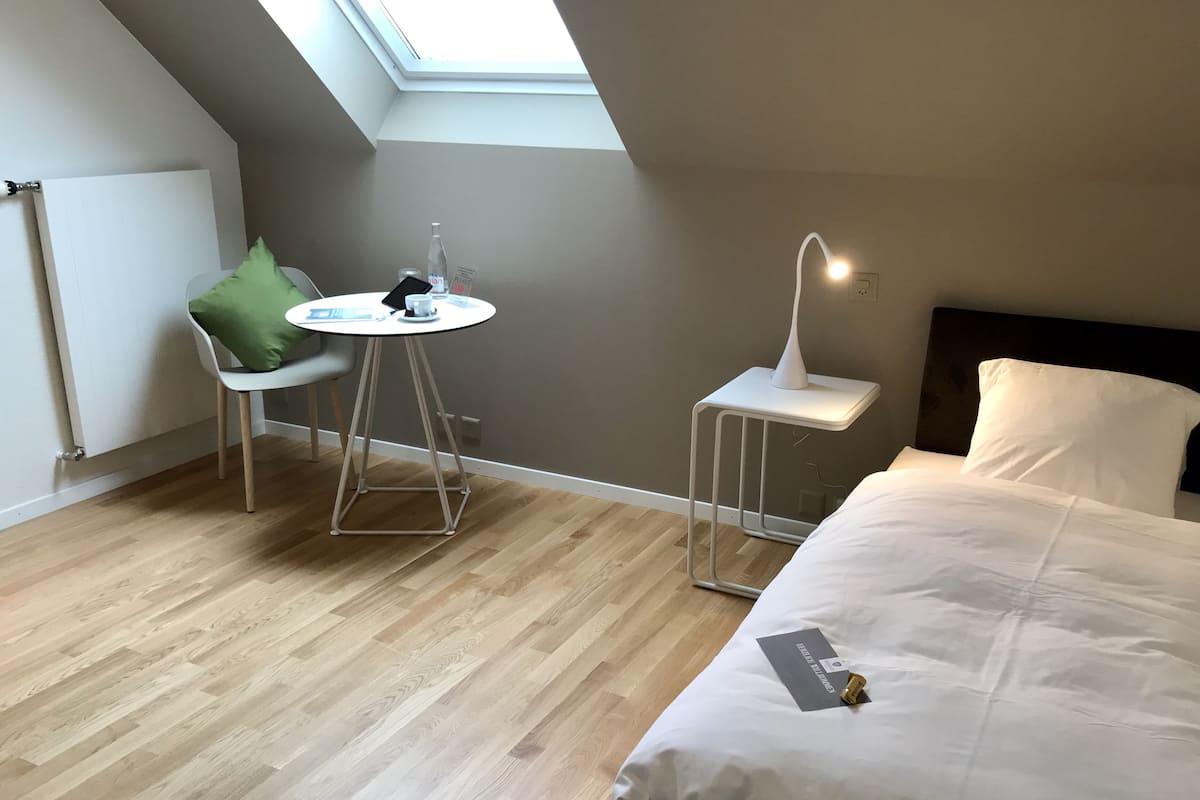 Una habitación de hotel moderna con tragaluz, una cama, una mesita redonda pequeña con una silla, una lámpara de noche y una pared blanca.