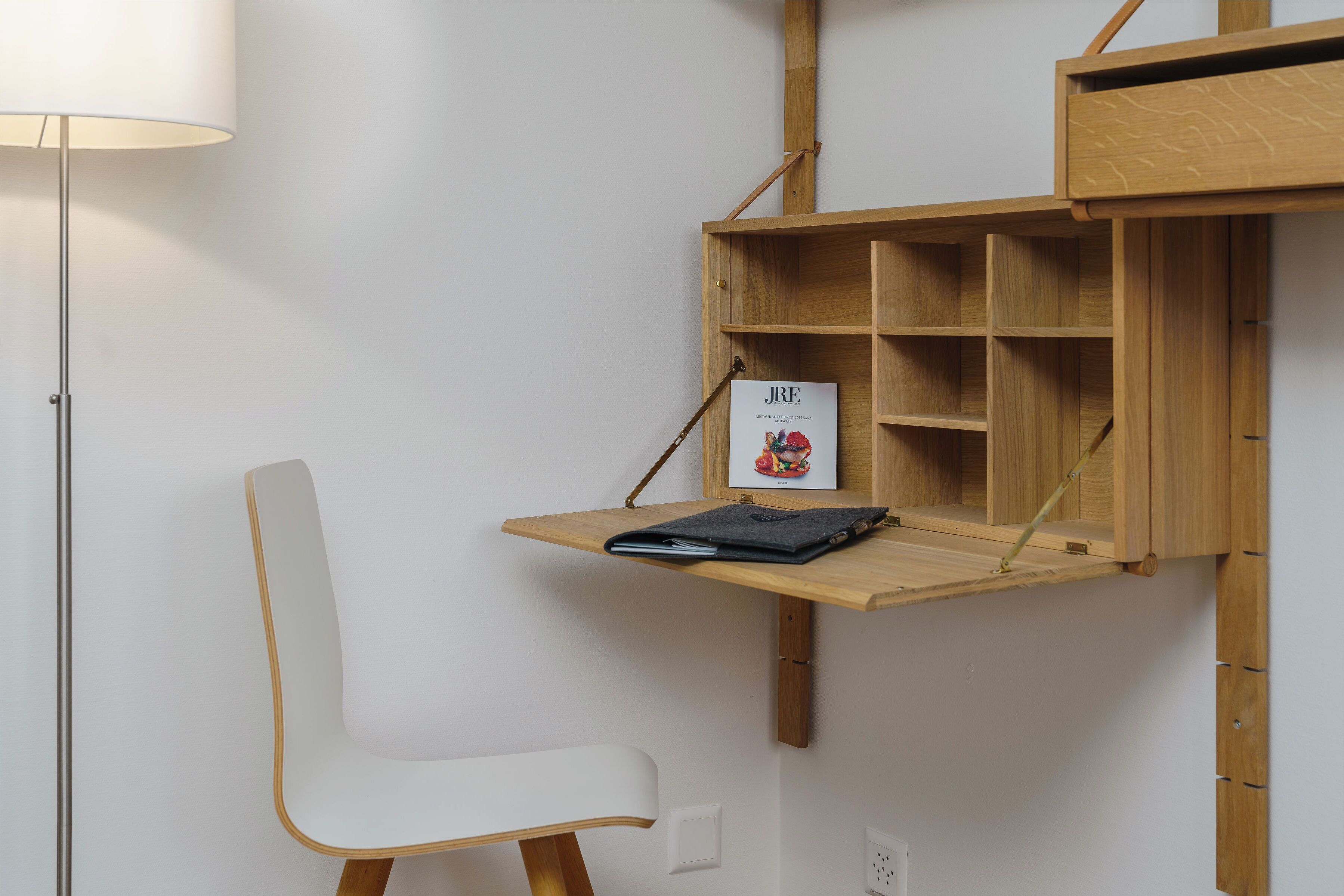 Ein schlichter Raum mit einem weißen Stuhl, einem Holztisch mit Ablagefläche und einer Lampe.