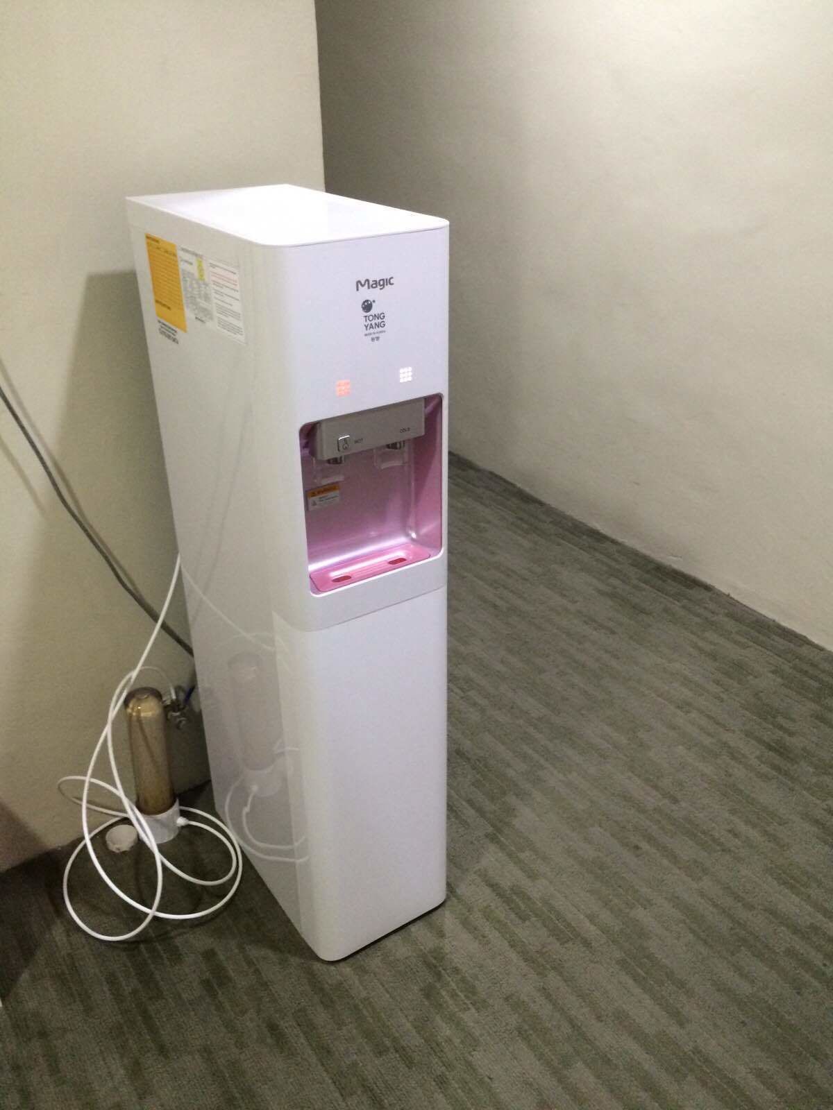 Ein weißer Wasserspender mit einer rosa Fontäne, platziert in einer Ecke mit grauem Teppichboden.