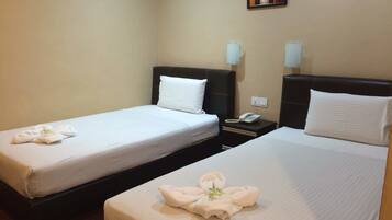 A hotel room with two beds, each with a white bedspread and a towel with a green center. There is a bedside table with a telephone and a wall-mounted light fixture.