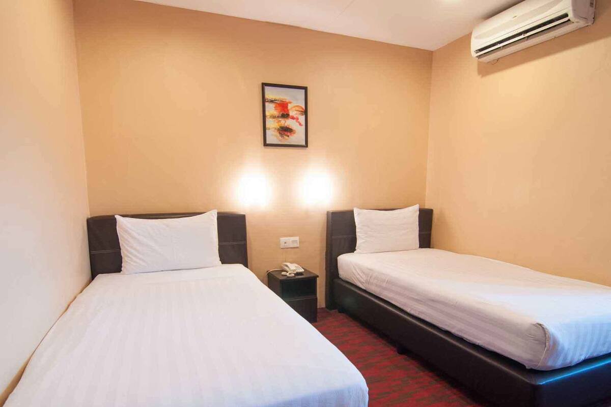 A hotel room with two single beds, a nightstand with a phone, and an air conditioning unit on the wall.