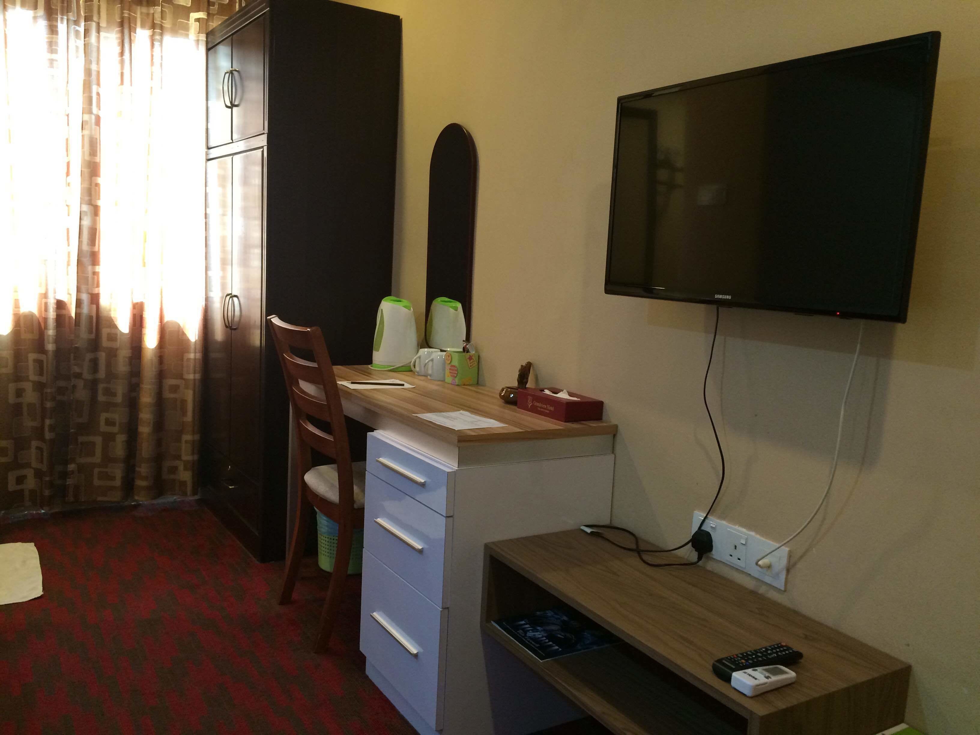 A hotel room with a desk, chair, television, and a small shelf with a remote control.