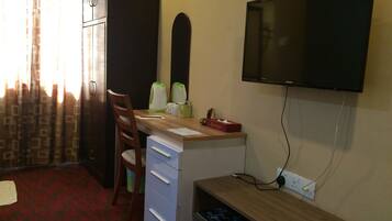 A hotel room with a desk, chair, television, and a small shelf with a remote control.