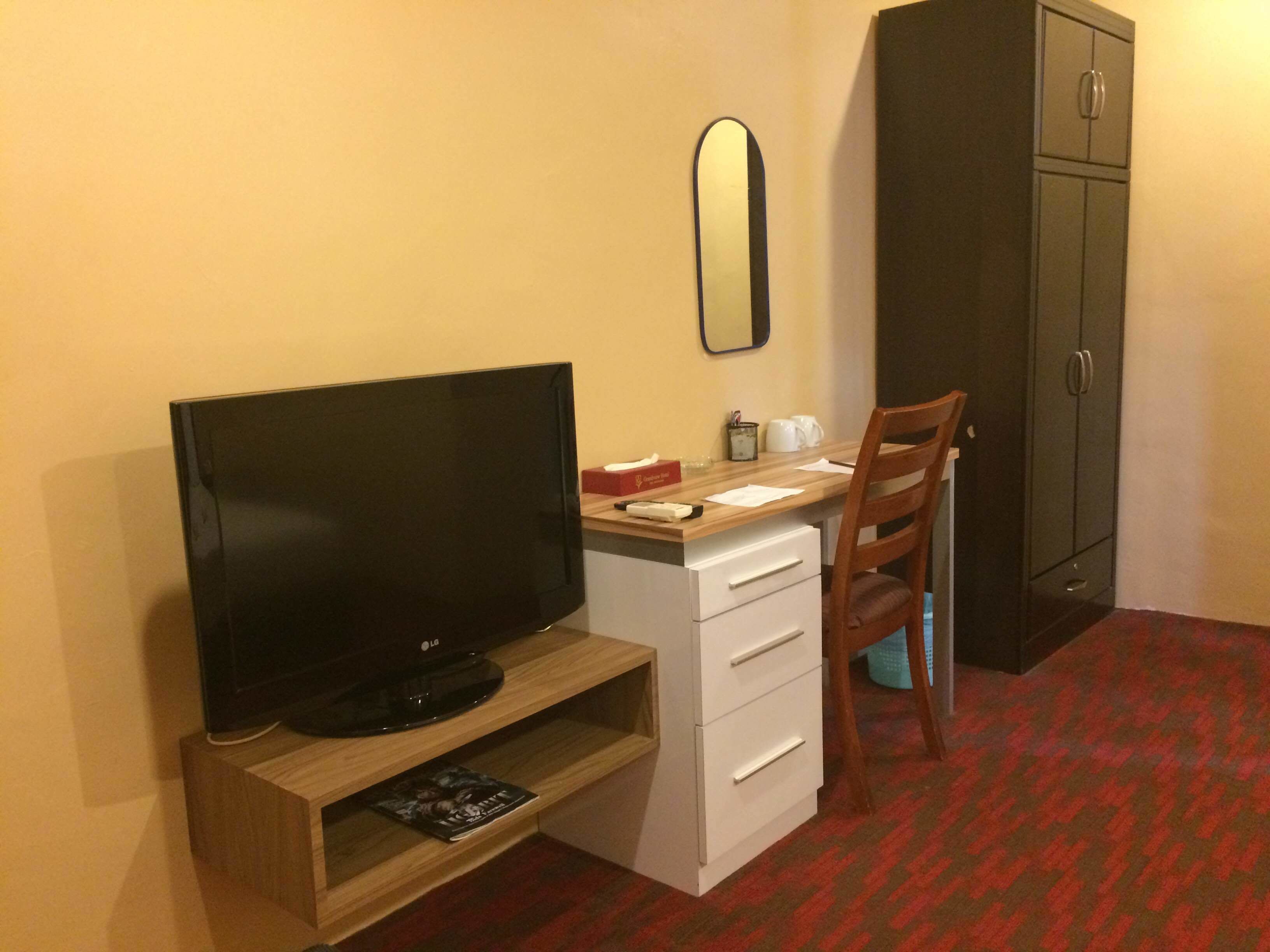 A hotel room with a TV on a wooden stand, a desk with a chair, a mirror, and a wardrobe.