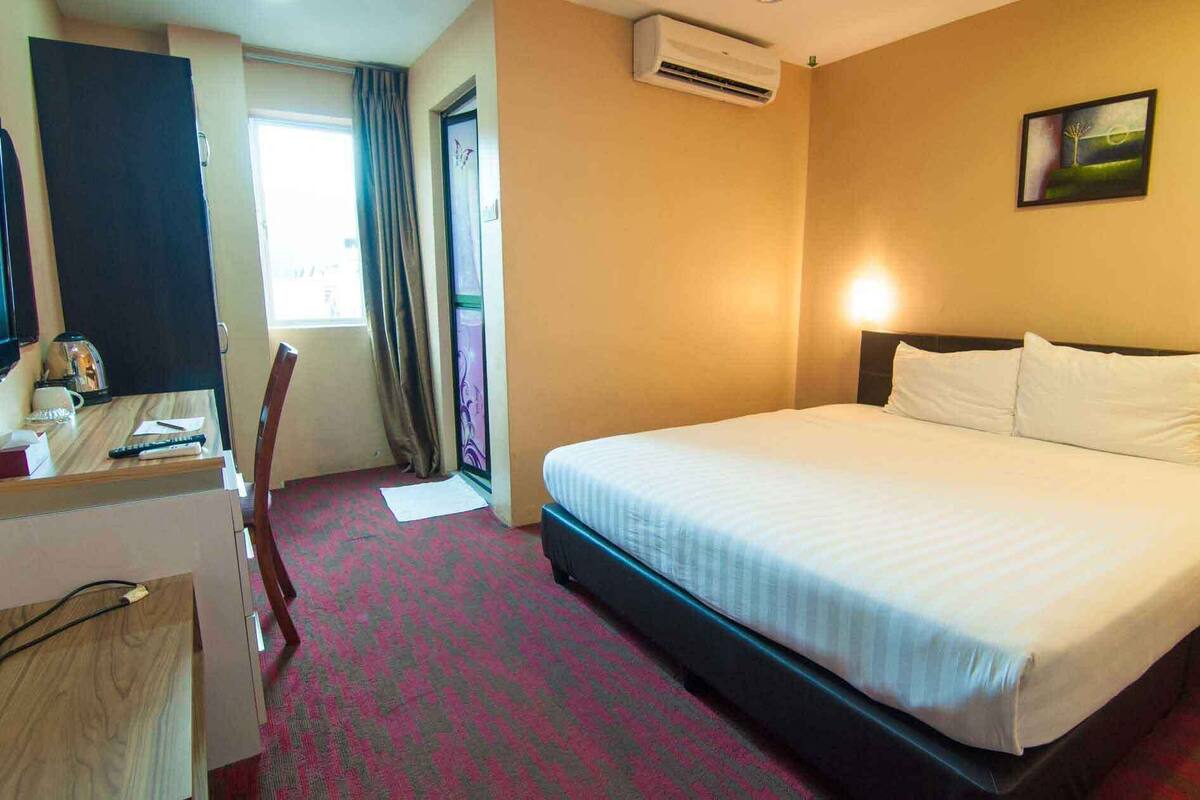 A hotel room with a bed, a desk, a chair, an air conditioning unit, and a painting on the wall.