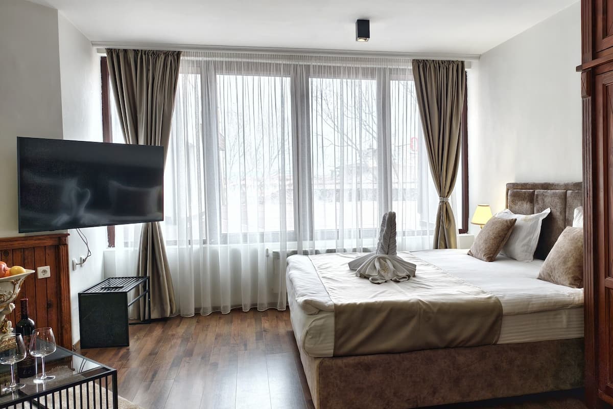 A hotel room with a large bed, a television, a wooden wardrobe, and a window with sheer curtains.