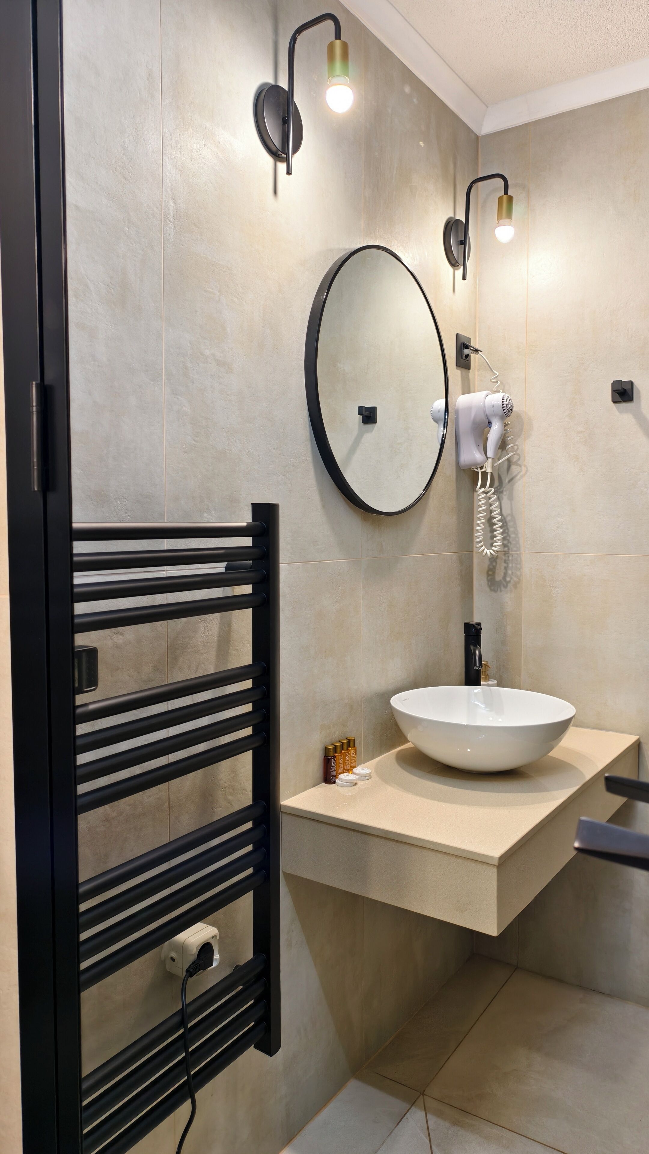 Une salle de bain moderne avec un porte-serviettes noir, un miroir rond, un lavabo blanc et deux appliques murales.