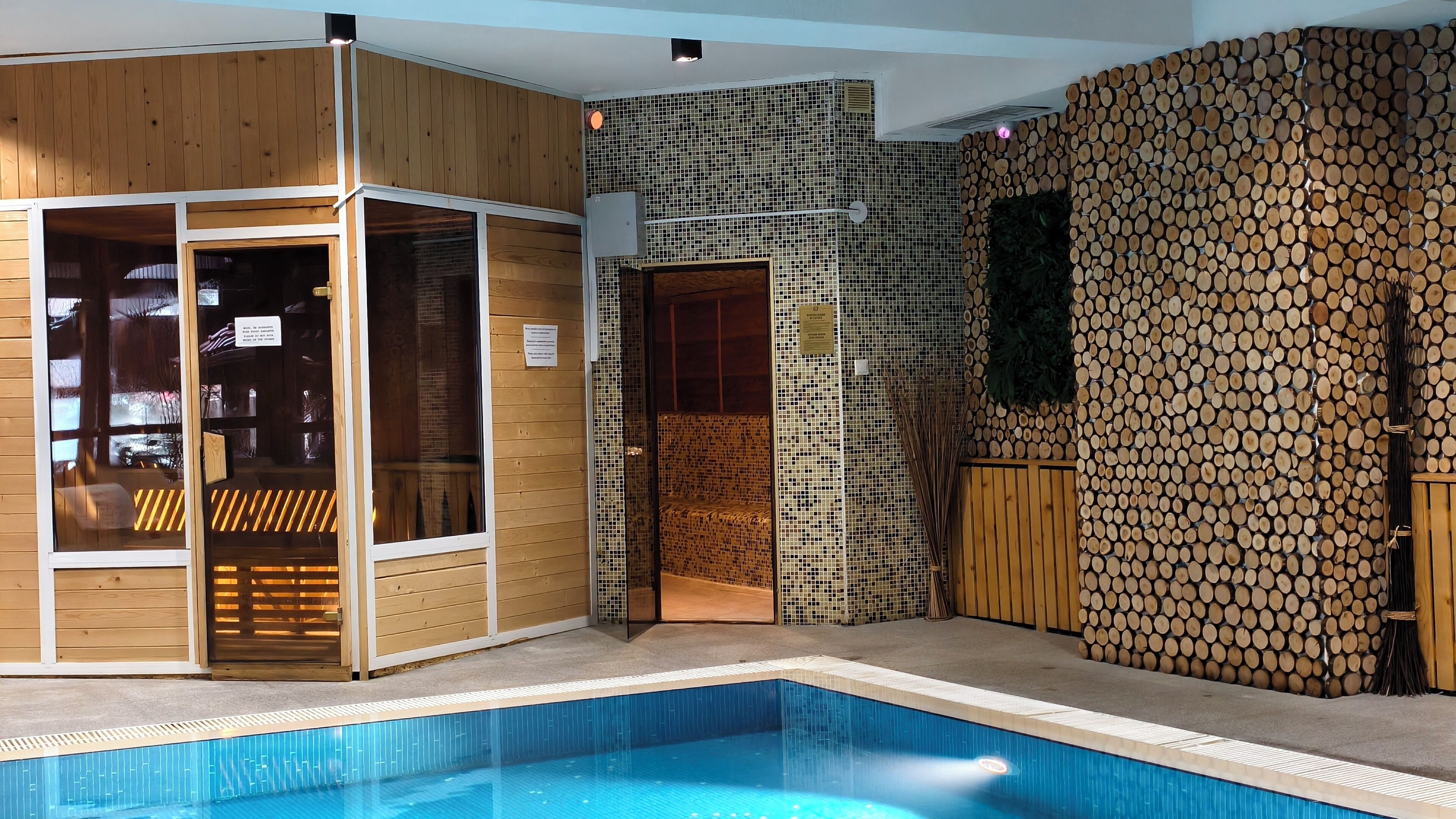 A modern indoor pool area with a wooden sauna, a glass-enclosed relaxation area, and a wall decorated with stacked logs.