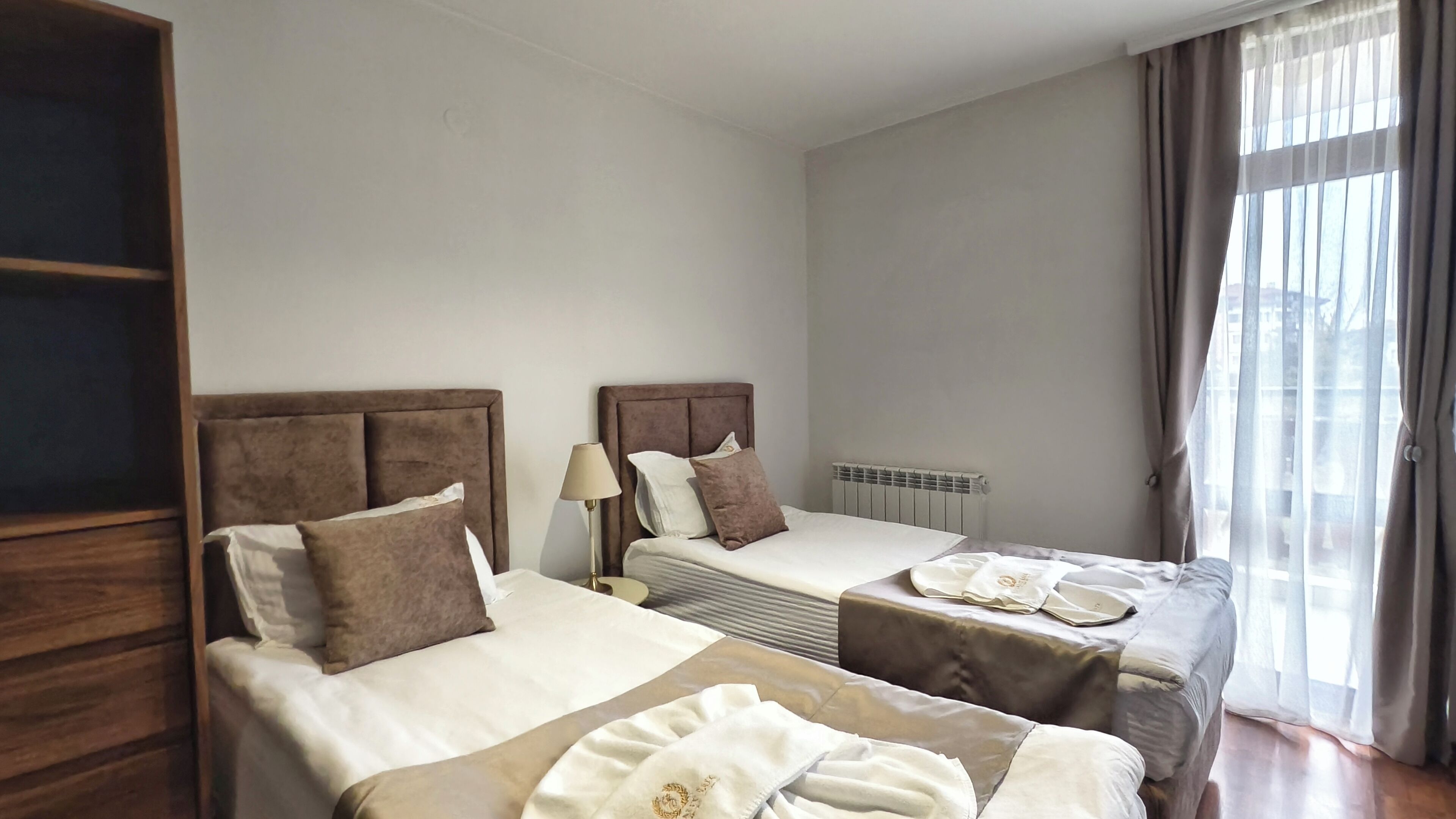 A hotel room with two beds, a wooden wardrobe, a radiator, and a window with curtains.