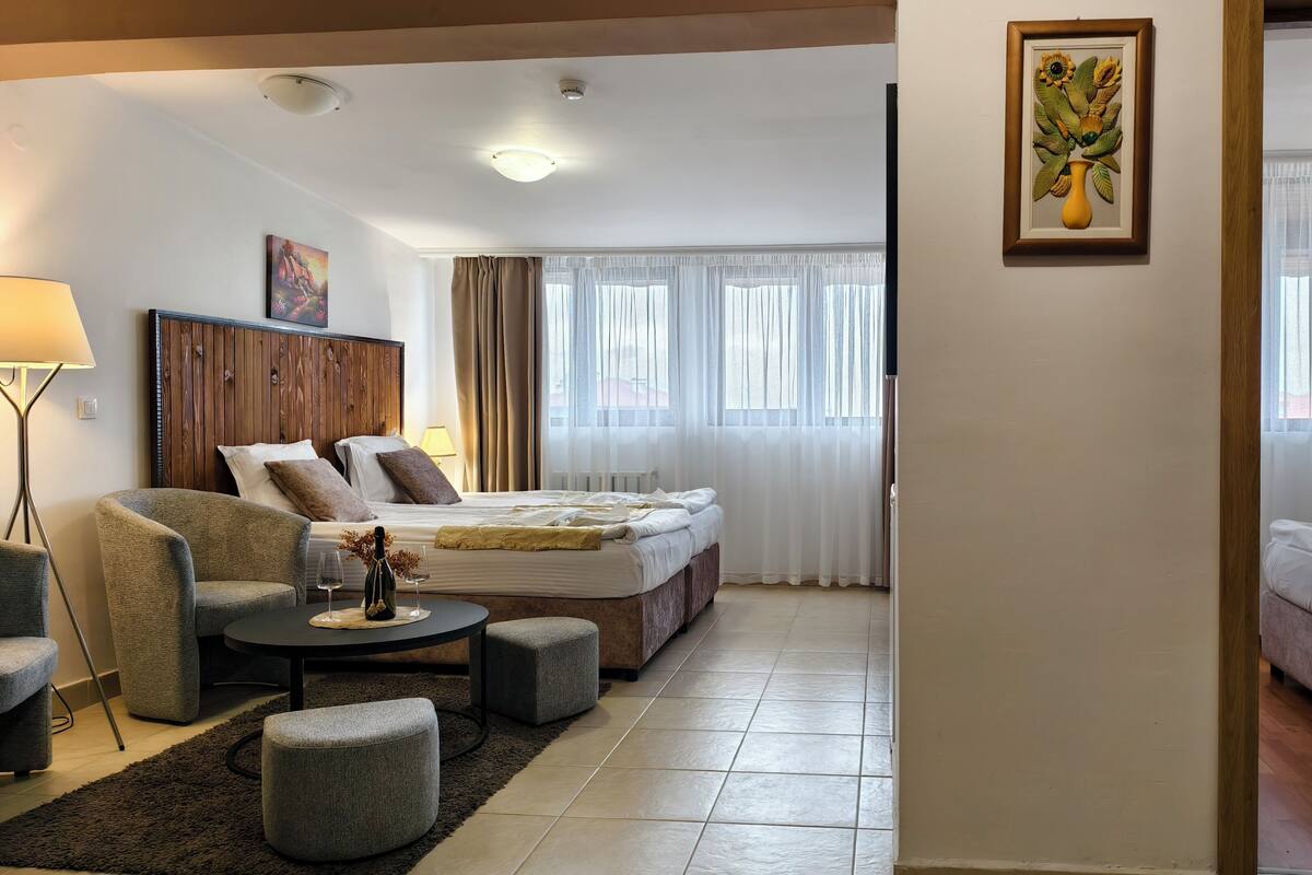 A hotel room with a bed, a sofa, a small table with a bottle and glasses, and a painting on the wall.