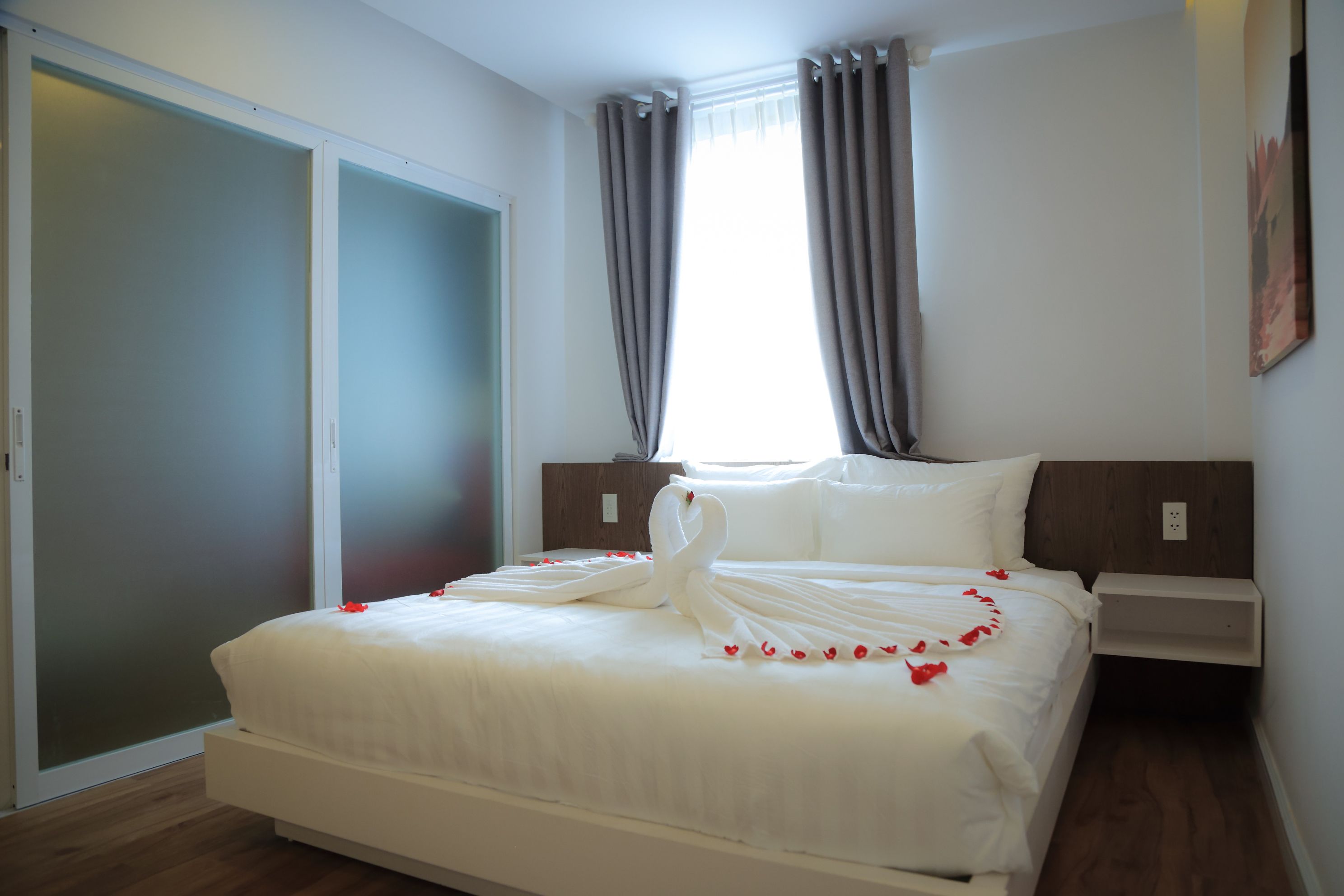 Hotelkamer met een bed, nachtkastje, houten vloer, schuifdeur en raam met gordijnen.