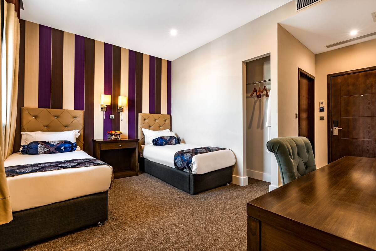 A hotel room with two beds, a wooden desk, a chair, a wardrobe, and a bathroom with a shower and towel rack.