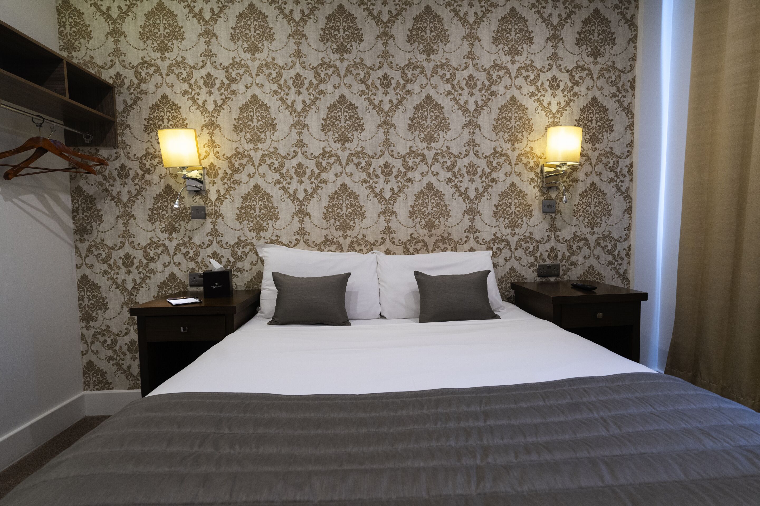 A neatly made bed with a dark grey bedspread and two pillows, flanked by nightstands with lamps. The room features ornate wallpaper and a wooden shelf.