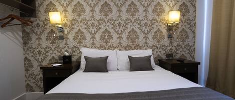 A neatly made bed with a dark grey bedspread and two pillows, flanked by nightstands with lamps. The room features ornate wallpaper and a wooden shelf.