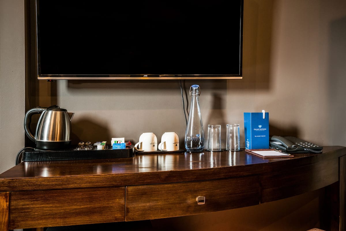 A hotel reception desk with a flat-screen TV, a coffee maker, cups, glasses, a phone, and a Grand Millennium brochure.