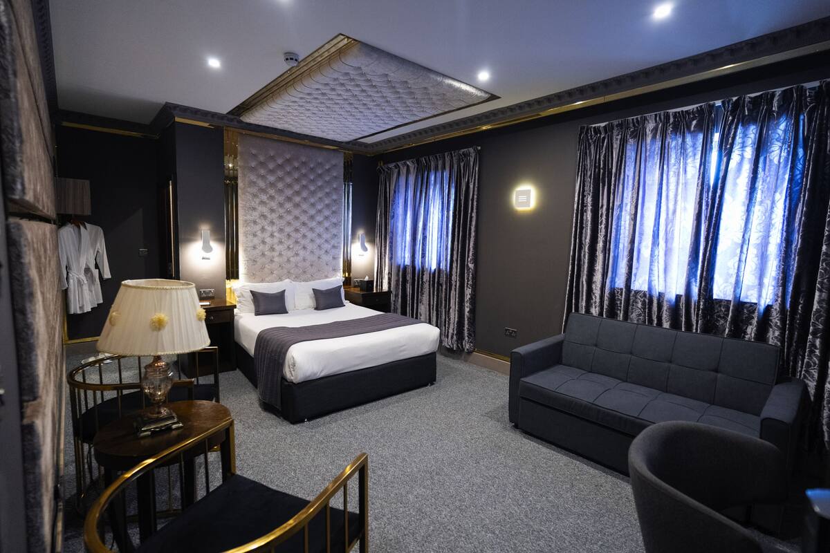 A modern hotel room with a large bed, a sofa, a desk with a lamp, and a wardrobe.