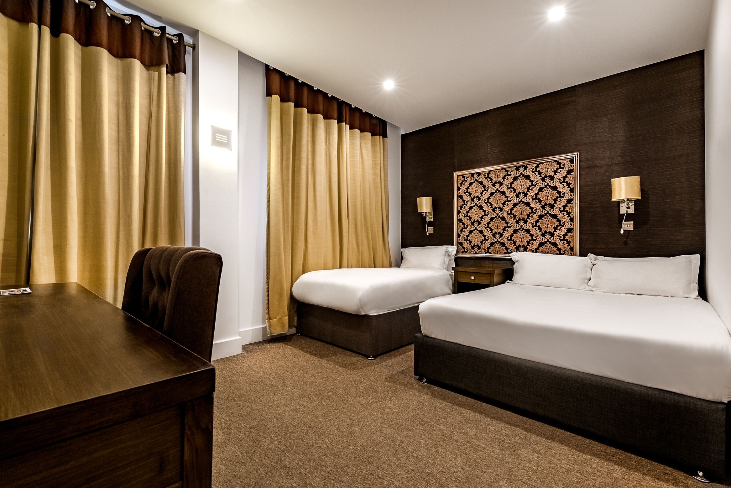 A hotel room with a wooden desk, two beds, a patterned headboard, and two wall-mounted lamps.