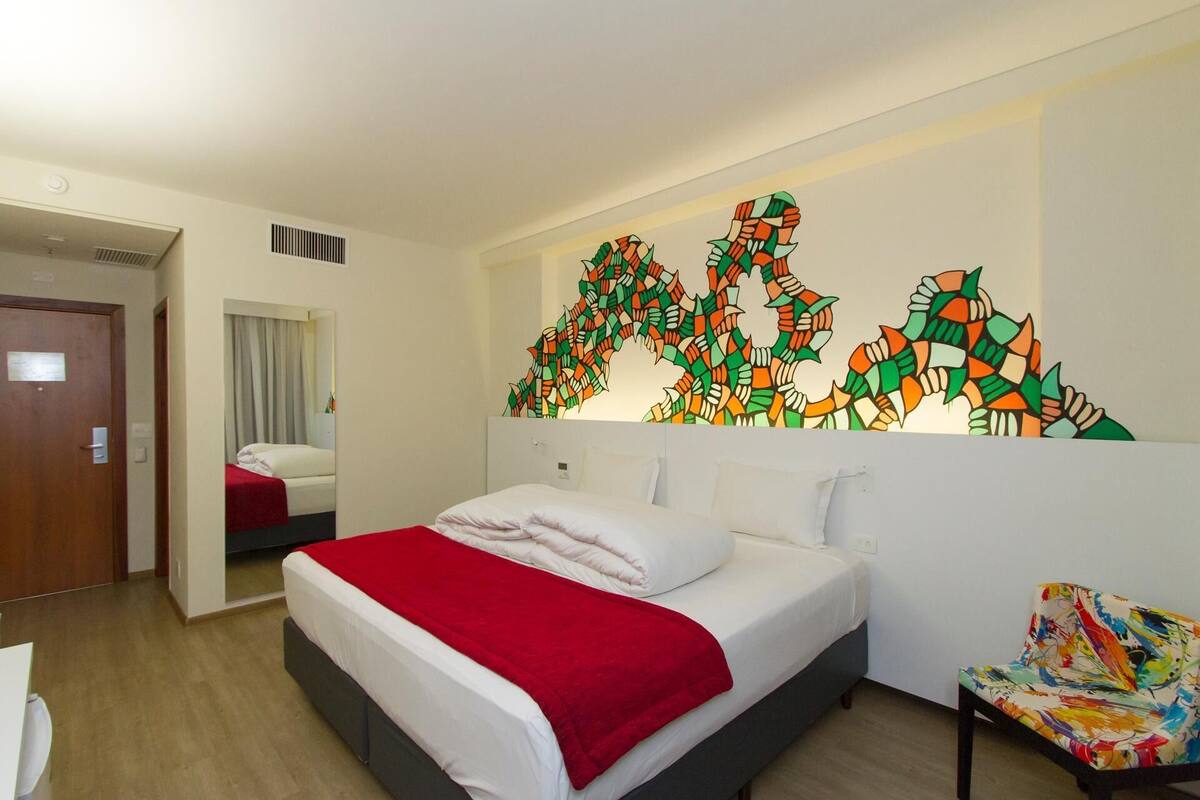 Een hotelkamer met een bed, een stoel, een spiegel en een muur met een kleurrijk abstract schilderij.