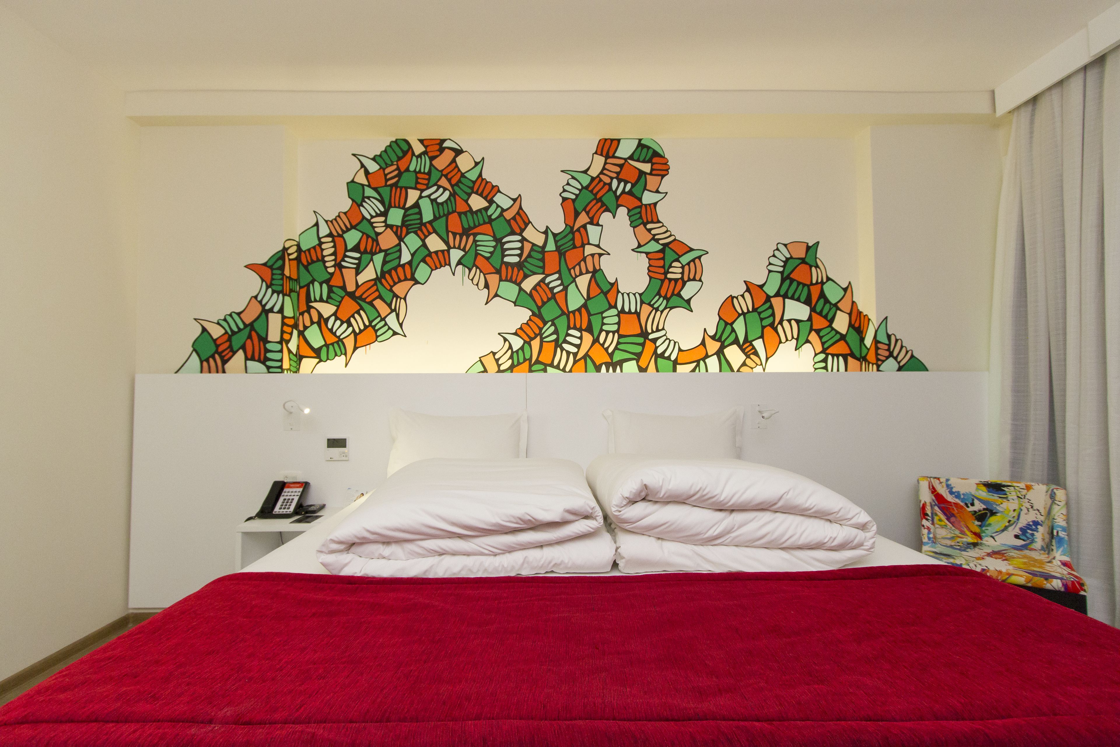 Quarto de hotel com cama, almofadas brancas, manta vermelha, mesa de cabeceira com telefone e arte abstrata colorida na parede.
