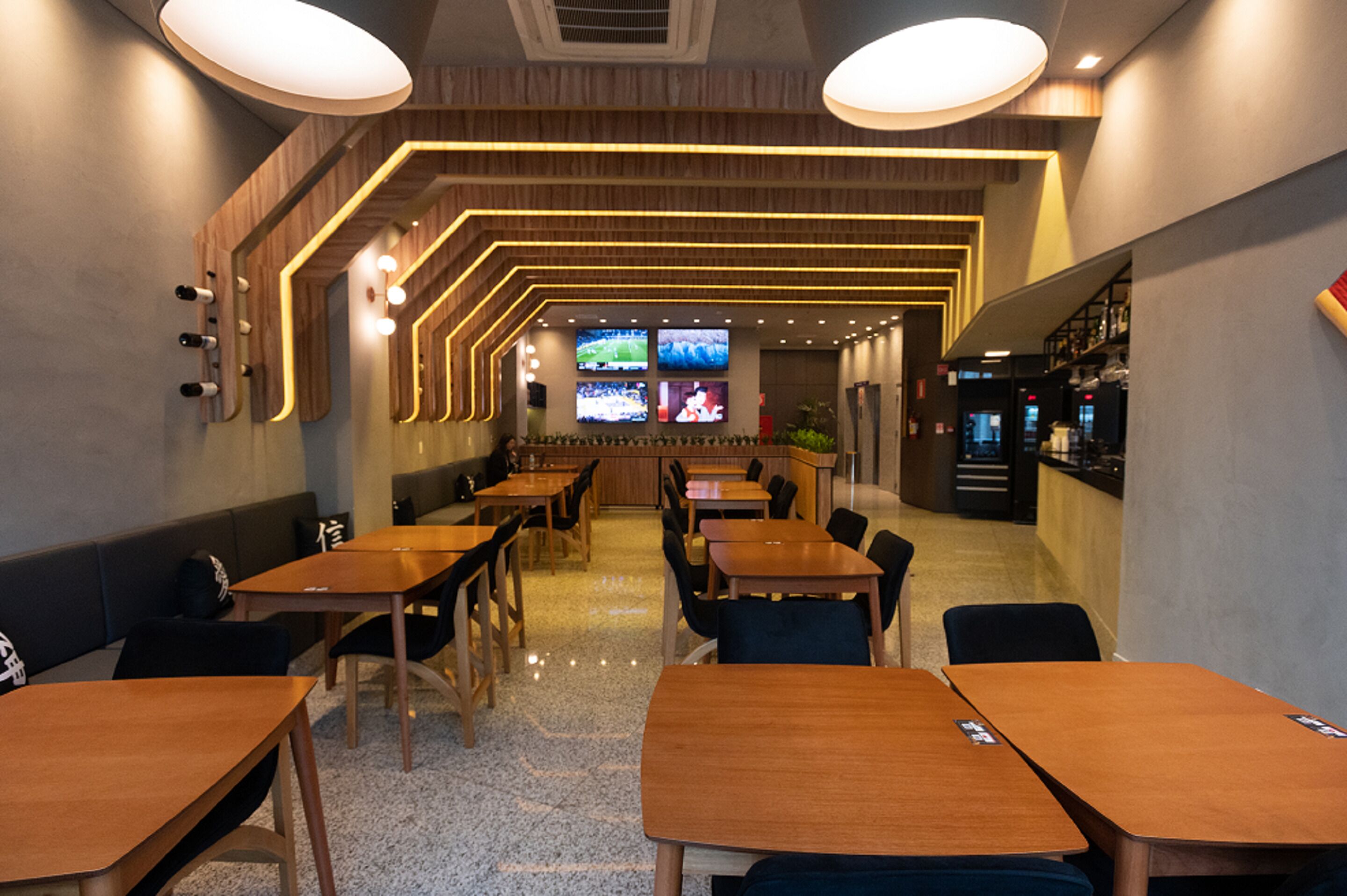A modern restaurant with wooden tables and chairs, a bar area, and multiple screens displaying content.
