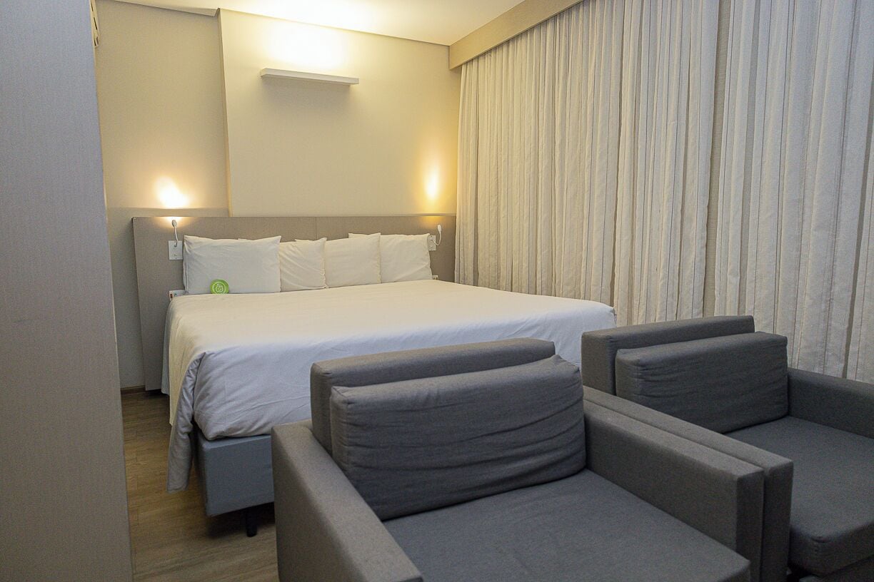 A hotel room with a bed, two grey armchairs, a bedside table, and a curtain.