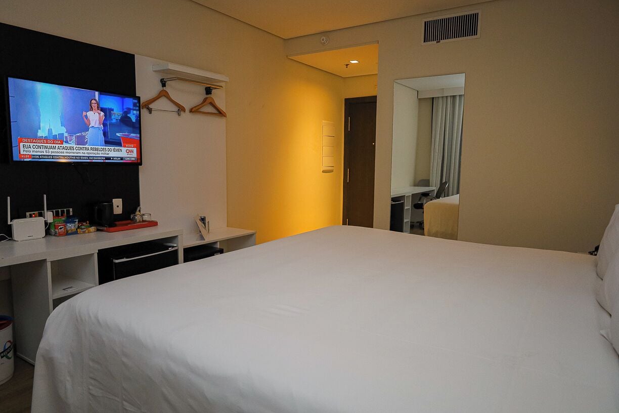 A hotel room with a bed, a TV mounted on the wall, a desk with a computer, and a mirror.