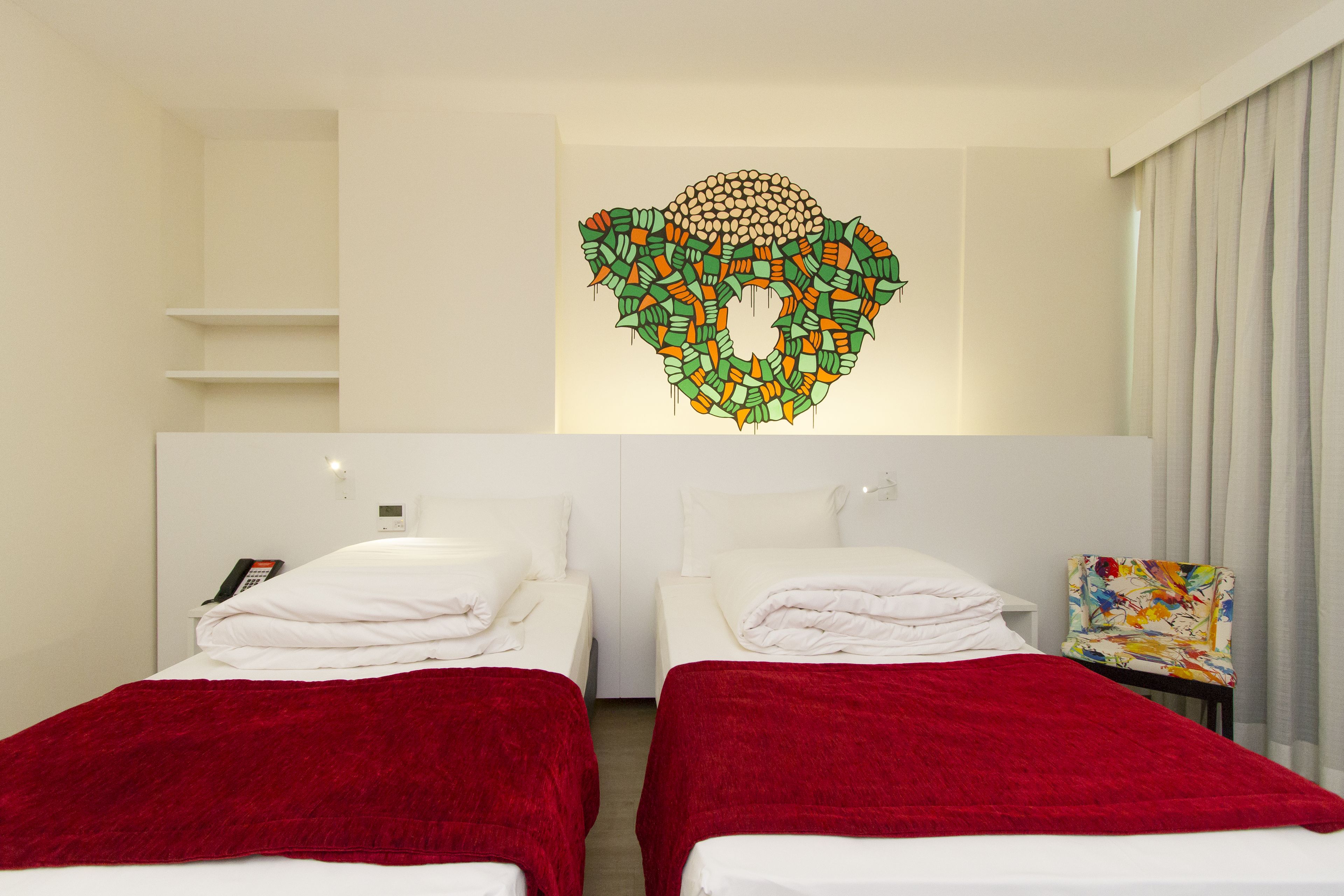 A hotel room with two beds, a red blanket on each, a colorful abstract artwork on the wall, and a chair with a floral pattern.