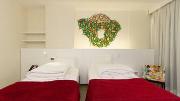 A hotel room with two beds, a red blanket on each, a colorful abstract artwork on the wall, and a chair with a floral pattern.