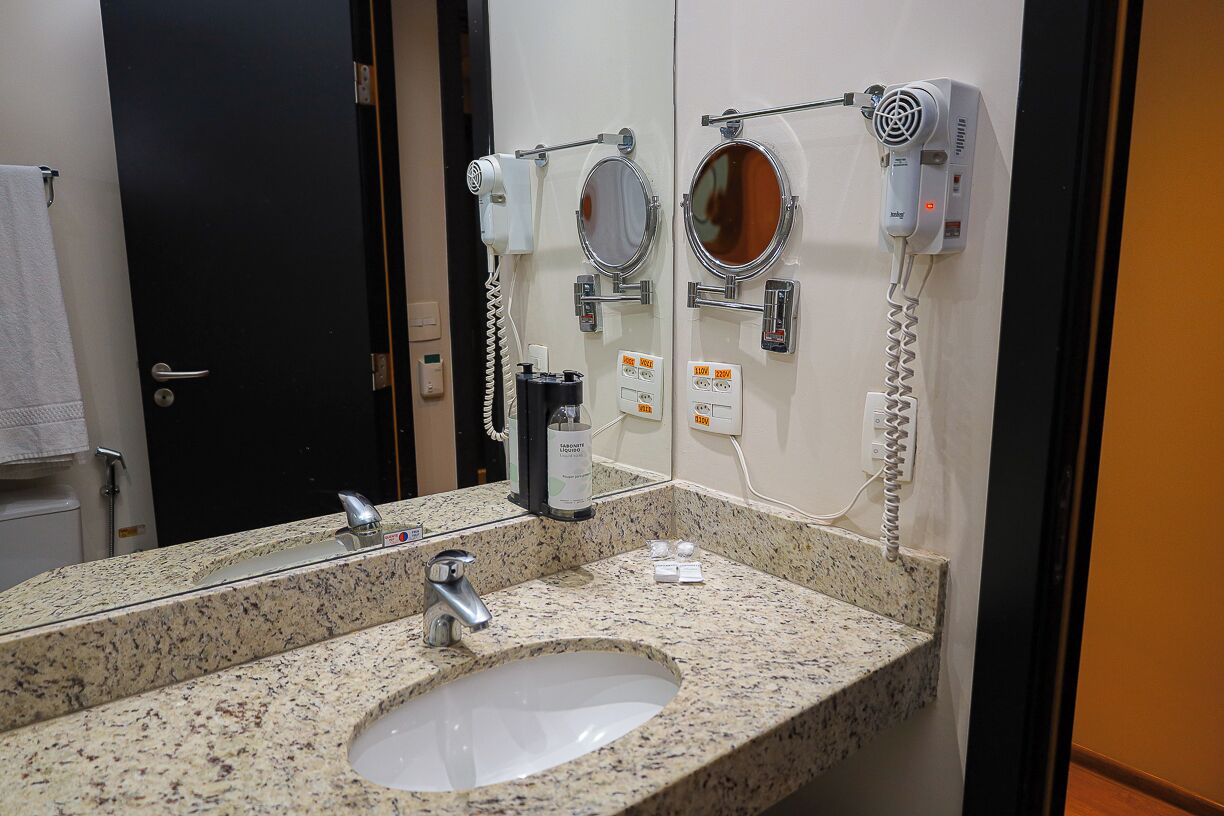 Casa de banho com lavatório com bancada de granito, um espelho e dois secadores de cabelo fixados na parede.