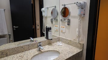 Casa de banho com lavatório com bancada de granito, um espelho e dois secadores de cabelo fixados na parede.