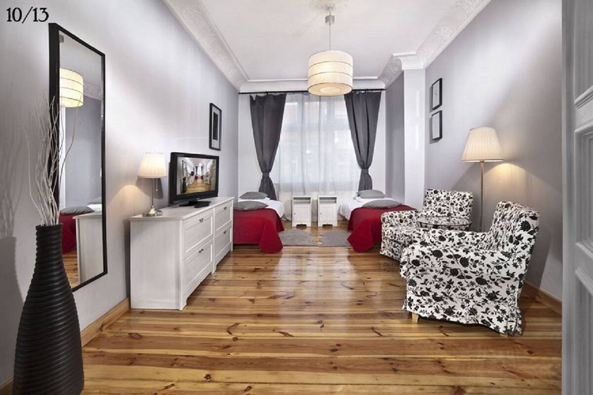 Une chambre d’hôtel moderne avec un plancher en bois, une commode blanche, une télévision, un lit avec une literie rouge, un canapé à motifs floraux et un grand miroir.