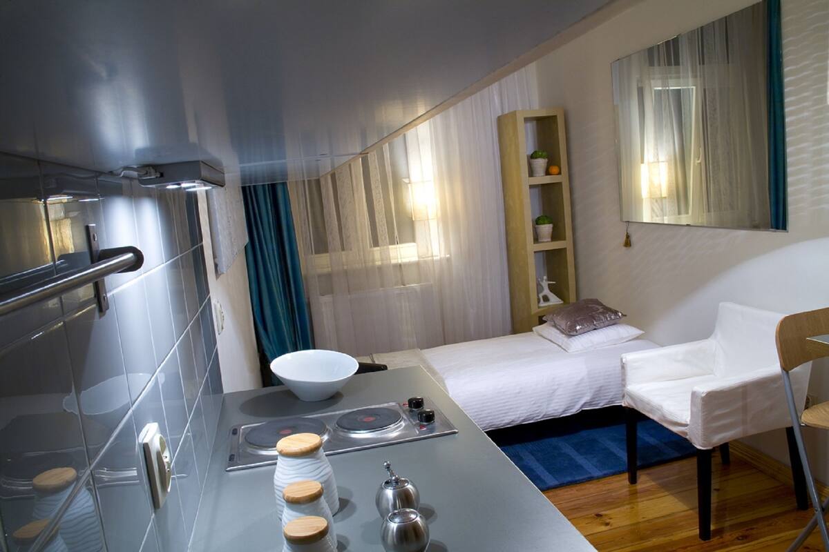 Une chambre d’hôtel moderne avec un lit, une chaise blanche, une petite table avec une bouilloire, un bol et une étagère ornée d’objets décoratifs.